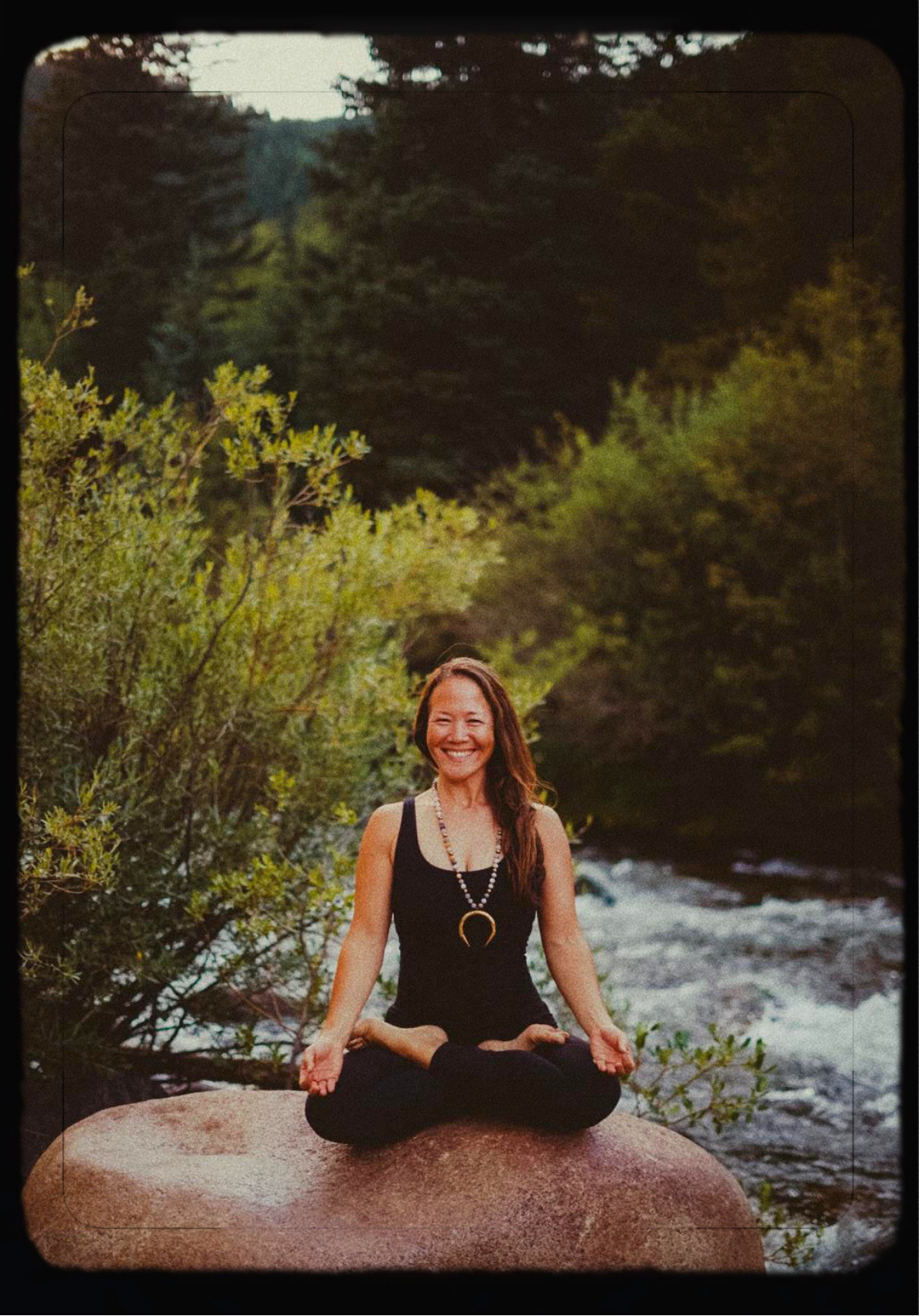 Mary Kirkendoll seated in lotus pose on a rock by a river